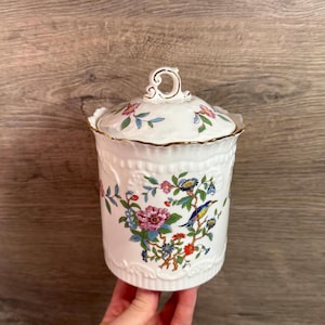 May include: A white ceramic jar with a lid, featuring a floral and bird design in pink, blue, and green. The jar has a gold rim and handle, and is held by a hand.