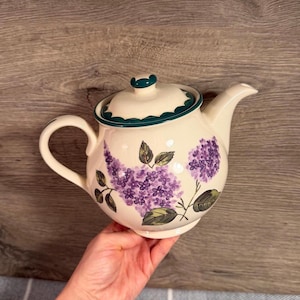 以下が含まれることがあります： 紫色のライラックの花房と緑の葉が手描きされた、クリーム色の陶磁器製ティーポット。ダークティール色の縁取りが施された蓋、曲線的なハンドル、先細りの注ぎ口が特徴で、全体に光沢のある釉薬で仕上げられています。