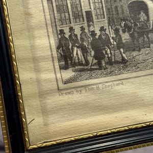 May include: An antique framed print depicting a street scene with figures and a horse-drawn carriage. The print, in black and white, features a building with the text "C TEMPLER WINE ME". The frame is black with a gold inner border.