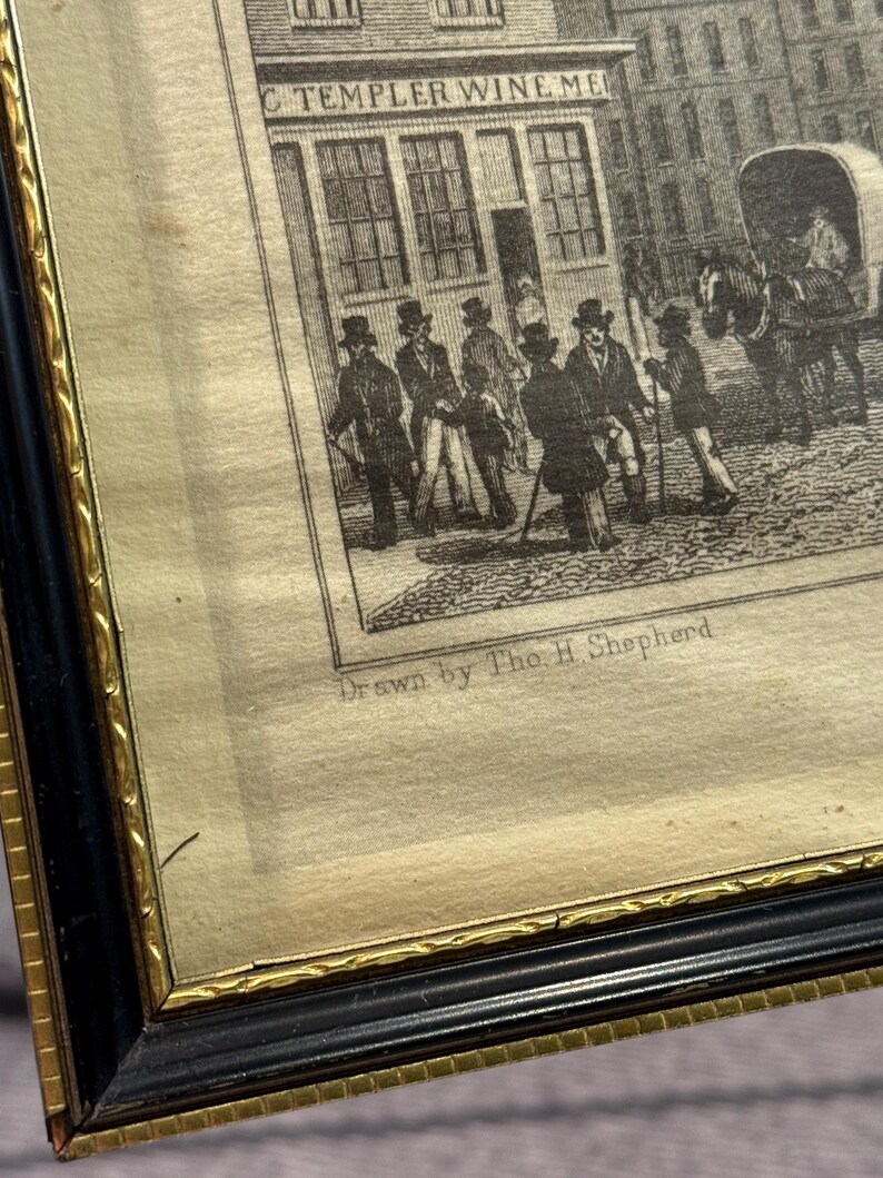 May include: An antique framed print depicting a street scene with figures and a horse-drawn carriage. The print, in black and white, features a building with the text "C TEMPLER WINE ME". The frame is black with a gold inner border.