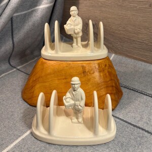 以下が含まれることがあります： バスケットを持った人の小さな人形が付いた白い陶器製のトーストラックが2つ。ラックは木製の台座の上に置かれています。