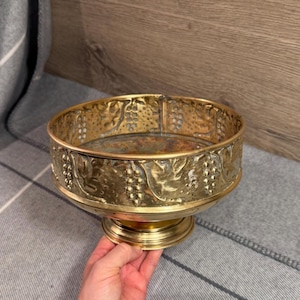 May include: A round brass bowl featuring an embossed design of grape clusters and vine leaves around the exterior. The bowl has a smooth, circular base and a polished metallic finish with visible texture in the relief pattern of the fruit.