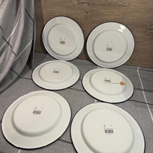 Pode incluir: Um conjunto de cinco pratos de esmalte branco com bordas pretas, vistos pela parte inferior. Cada prato apresenta uma base circular central, um acabamento liso e brilhante e uma pequena etiqueta de c&oacute;digo de barras com o texto AT-EN202353.