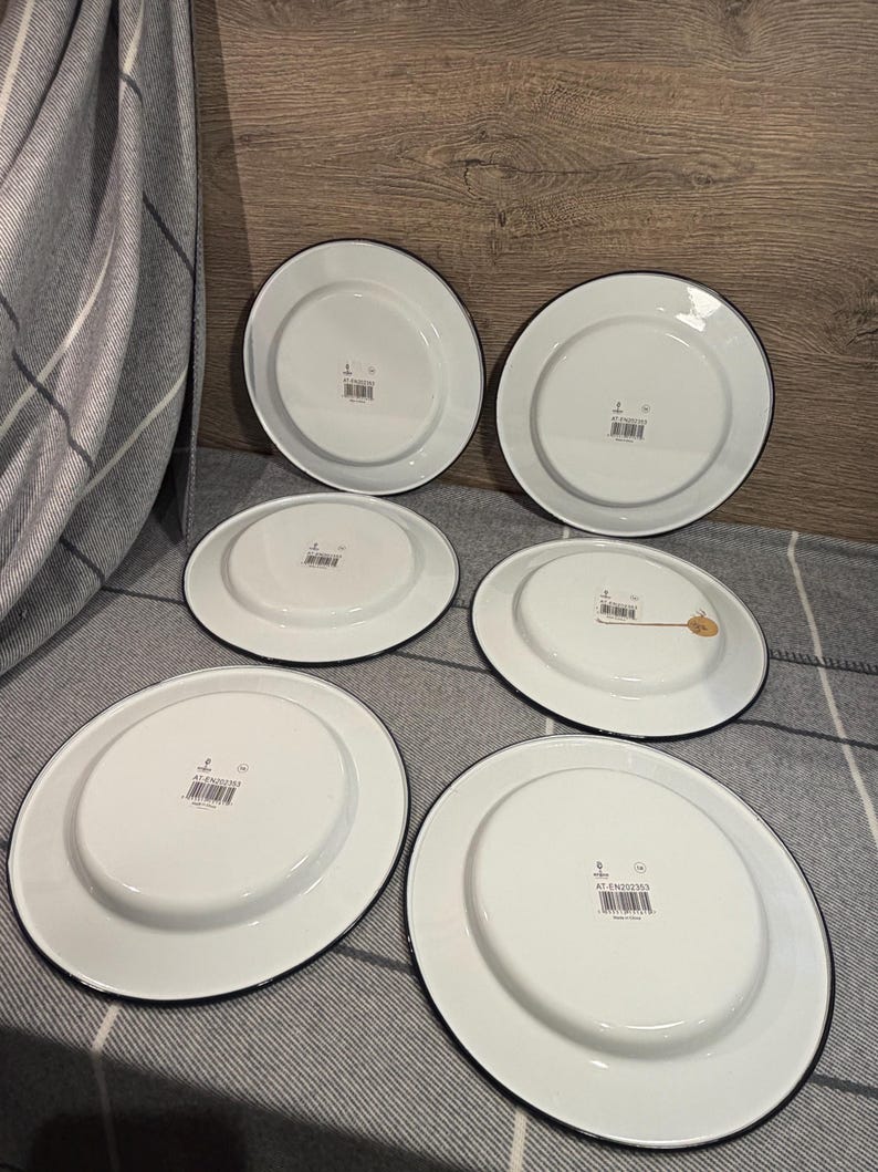 Pode incluir: Um conjunto de cinco pratos de esmalte branco com bordas pretas, vistos pela parte inferior. Cada prato apresenta uma base circular central, um acabamento liso e brilhante e uma pequena etiqueta de c&oacute;digo de barras com o texto AT-EN202353.