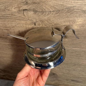 May include: A clear glass condiment jar encased in a polished chrome metal frame. The container features a hinged circular lid with a small attached spoon and a curved handle extending from the side, all finished in a reflective silver tone.