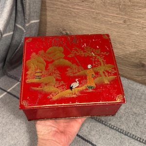 Peut inclure: Une boîte en métal rouge avec un motif de paysage asiatique doré. La boîte représente une personne en robe, une grue, des arbres et une pagode. La boîte est de forme carrée et possède une bordure dorée.