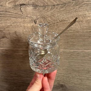 Puede incluir: Un azucarero transparente de cristal tallado con tapa y una cucharita. El cuenco tiene un diseño de diamantes y un pomo en la tapa. La cuchara es plateada. El azucarero se sostiene en una mano.