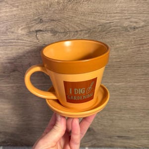 May include: Orange ceramic mug and saucer set, designed to resemble a flower pot. The mug has a handle and the text "I DIG GARDENING" with a shovel graphic. The set is a warm, earthy tone.