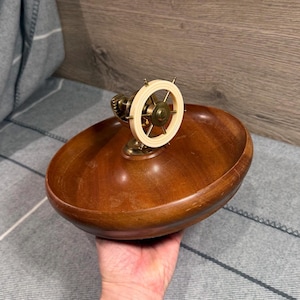 Può includere: Una ciotola rotonda in legno dotata di un meccanismo centrale in ottone sormontato da un timone di nave color crema. La ciotola presenta una finitura liscia e lucida con venature del legno visibili, mentre il pezzo centrale metallico include un ingranaggio.