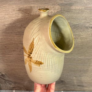 Puede incluir: Una salera de cerámica con un esmalte marrón y blanco. La salera tiene una forma única con una gran abertura en el lateral. La abertura está diseñada para permitir un fácil acceso a la sal.