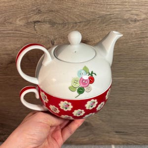 Puede incluir: Tetera blanca con un patrón floral rojo y blanco y una tapa. La tetera tiene un infusor incorporado y una taza a juego.