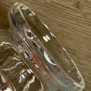 Op de afbeelding: Een helder glazen object met een brede, gebogen rand. Het glas heeft een getextureerd oppervlak en reflecteert licht, waardoor highlights en schaduwen ontstaan. Het object lijkt een decoratief stuk of een deel van een groter vat te zijn.