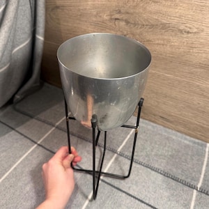 May include: A silver metal planter with a rounded bowl shape, supported by a black metal stand. The planter is empty and appears to be made of a brushed metal. The stand has a simple, geometric design. The background is a grey and white striped blanket.
