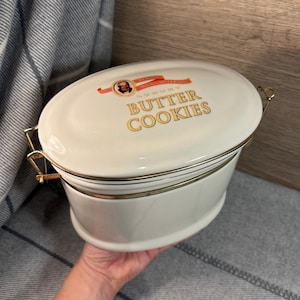 Puede incluir: Tarro de galletas de cerámica blanca con tapa y ribete dorados. La tapa tiene una cinta dorada con el texto "Mrs. McWatters Luxury Butter Cookies".