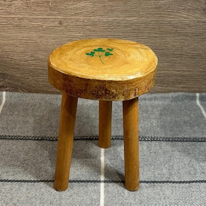 May include: A small, wooden stool with three legs. The round seat is light brown with a green clover design. The legs are a darker shade of brown. The stool is approximately 25 cm tall.