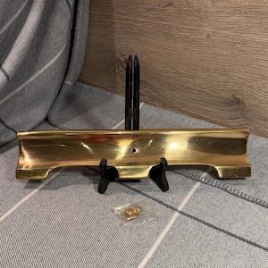 May include: A polished gold-tone metal curved tray with a central circular hole, supported by a black plastic stand. A small, separate gold-tone metal knob is enclosed in a clear plastic bag positioned in front of the tray on the surface.