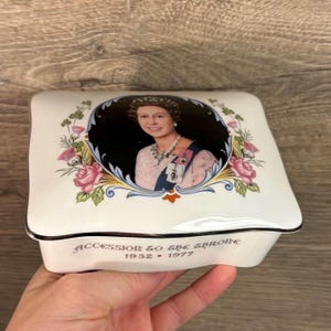 Peut inclure: Boîte en porcelaine blanche avec une bordure argentée et une image de la reine Élisabeth II. La boîte porte l'inscription "Accession to the Throne 1952 - 1977" sur le fond.