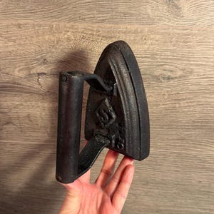 Puede incluir: Una plancha antigua negra con asa. La plancha tiene forma triangular con un extremo puntiagudo y un emblema decorativo. El asa es cilíndrica y está unida al cuerpo de la plancha. La plancha se sostiene en una mano.