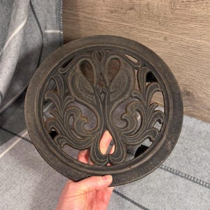 May include: A round, cast iron trivet with a decorative design featuring two stylized birds facing each other, surrounded by ornate scrollwork. The trivet has a dark, aged finish and is designed to protect surfaces from hot cookware.