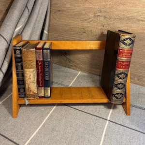May include: A wooden bookshelf with five books on the top shelf and one book on the bottom shelf. The books are all antique and have gold lettering on the spines. The top shelf books are titled "The Lair of the White Worm", "Far from the Madding Crowd", and "Jane Eyre". The bottom shelf book is titled "Cyclopaedia of English Literature, Chambers, Vol II".
