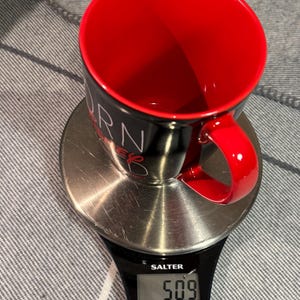 Peut inclure: Une tasse &agrave; caf&eacute; noire et rouge avec le mot "BURN" visible, pos&eacute;e sur une balance num&eacute;rique. La balance affiche un poids de 509, probablement en grammes. La tasse a un int&eacute;rieur et une anse rouges. La balance est de marque Salter.