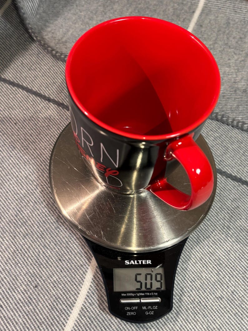 Peut inclure: Une tasse &agrave; caf&eacute; noire et rouge avec le mot "BURN" visible, pos&eacute;e sur une balance num&eacute;rique. La balance affiche un poids de 509, probablement en grammes. La tasse a un int&eacute;rieur et une anse rouges. La balance est de marque Salter.