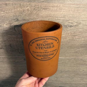 May include: A cylindrical, terracotta-colored kitchen utensil holder. The holder features a black oval label with the text "THE ORIGINAL SUFFOLK JAR KITCHEN UTENSILS" and "HENRY WATSON POTTERY ENGLAND Founded 1800".