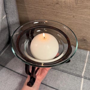 May include: A lit white spherical candle sits inside a clear glass bowl. The bowl is held by a dark brown metal stand with decorative scrollwork. The candle holder is a decorative home accent.