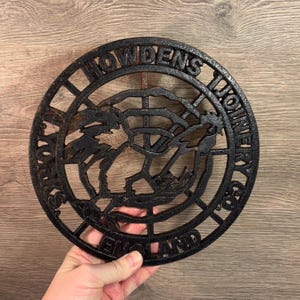 Könnte beinhalten: Schwarzer gusseiserner Untersetzer mit den Worten "Howdens Ironmongery Co. England" am Rand. Die Mitte des Untersetzers zeigt ein stilisiertes Design eines Hahns.