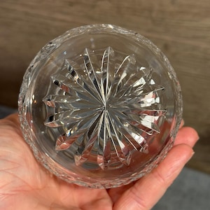 Op de afbeelding: Heldere kristallen glazen kom met een stervormig ontwerp op de basis. De kom heeft een gesneden patroon rond de rand. De kom wordt in de hand van een persoon gehouden.