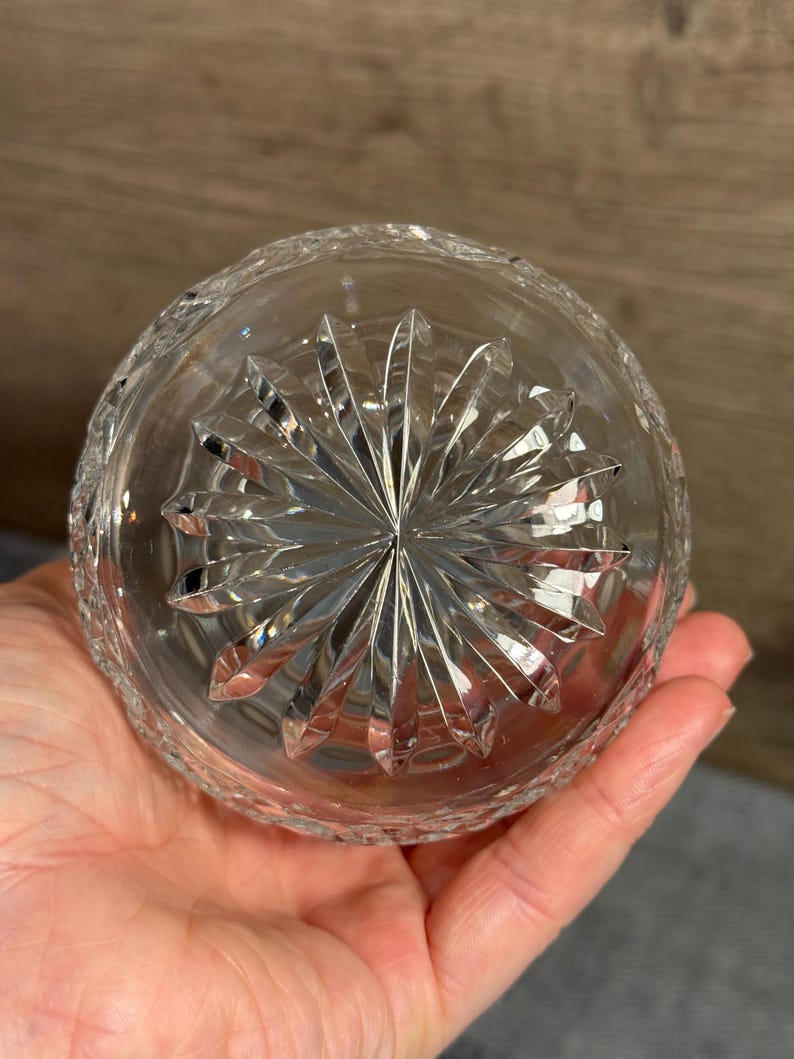 Op de afbeelding: Heldere kristallen glazen kom met een stervormig ontwerp op de basis. De kom heeft een gesneden patroon rond de rand. De kom wordt in de hand van een persoon gehouden.