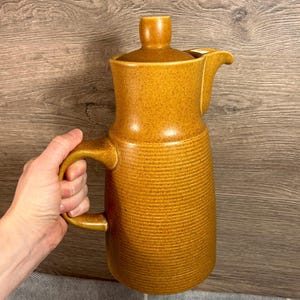 Peut inclure: Une cruche en céramique de couleur moutarde avec un couvercle et une poignée. La cruche présente un motif texturé et nervuré sur le corps et un bec verseur. Le couvercle a un petit bouton sur le dessus. La cruche est tenue par une main.
