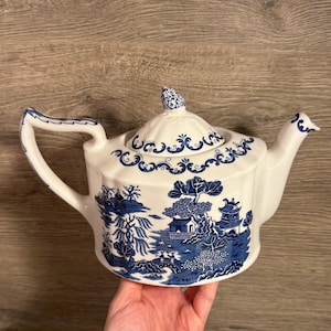 May include: White ceramic teapot with a blue Willow pattern design. The teapot features a curved handle, a spout, and a lid with a decorative finial. The blue and white design depicts a landscape scene with trees, buildings, and a bridge.