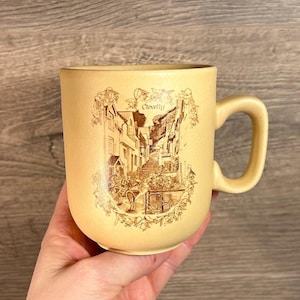 May include: A beige ceramic mug featuring a brown illustration of a street scene in Clovelly, England. The mug has a handle and is decorated with floral designs.