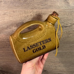 以下が含まれることがあります： 黄色のロープの取っ手が付いた茶色の陶器の壺。壺には「Lassetters Gold」という文字が書かれています。