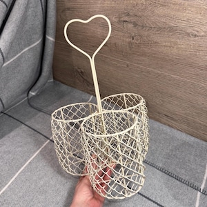 May include: Off-white wire utensil holder with a heart-shaped handle. The holder has three cylindrical compartments with a diamond-patterned mesh design. The holder is on a gray and white plaid blanket.