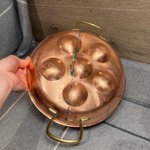 May include: A copper escargot pan with six round indentations. The pan has two brass handles and a crack with turquoise paint. The pan is round and has a polished finish. The pan is sitting on a gray and white striped surface.