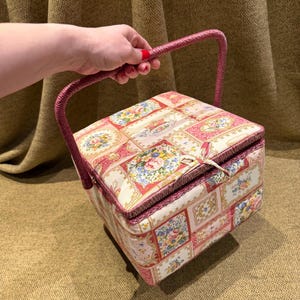 Può includere: Un cestino da cucito vintage con manico rosa e motivo floreale. Il cestino quadrato presenta un coperchio incernierato ed è decorato con un motivo ripetuto di bouquet floreali e bordi decorativi nei toni del rosa, bianco e blu. Il manico è rosa abbinato.