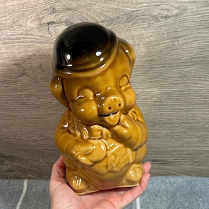 May include: A brown ceramic piggy bank with a black hat. The piggy bank is smiling and has its tongue sticking out. The piggy bank is holding its hand to its mouth.