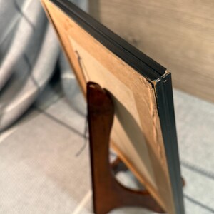 May include: A framed artwork with a black frame is displayed on a wooden easel. The artwork is visible from the side, revealing a tan backing and a glimpse of the image within the frame. The easel is a dark brown color.