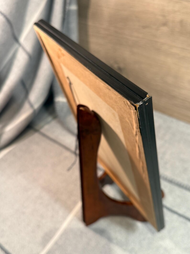 May include: A framed artwork with a black frame is displayed on a wooden easel. The artwork is visible from the side, revealing a tan backing and a glimpse of the image within the frame. The easel is a dark brown color.