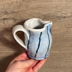 May include: A small, handmade ceramic pitcher with a white handle and rim. The body is light blue with dark blue vertical stripes. The pitcher is held in a hand against a wooden background.