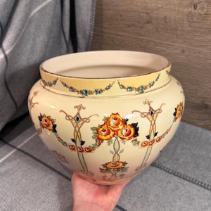 Op de afbeelding: Een crèmekleurige keramische kom met een brede opening en een ronde vorm. De buitenkant is versierd met een herhalend patroon van oranje rozen, groen blad en dunne goudkleurige lijnen, met een bloemenslinger langs de rand van de kom.