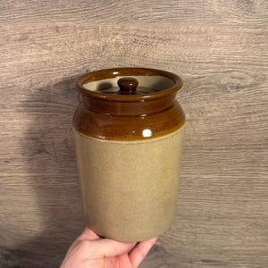 Może przedstawiać: Wazon z kamionki w stylu vintage z pokrywką. Wazon ma cylindryczny kształt z jasnobrązowym korpusem oraz ciemnobrązową obręczą i pokrywką. Pokrywka ma mały, okrągły uchwyt. Wazon jest prawdopodobnie używany do przechowywania żywności.