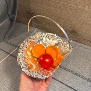 May include: A silver-toned metal basket with a decorative embossed floral rim and a thin arched handle. The basket contains two round orange fruits and one round red fruit, all resting inside the reflective, scalloped-edge bowl structure.