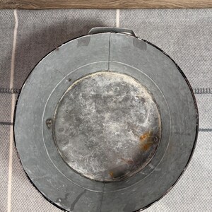 Op de afbeelding: Een ronde bak van gegalvaniseerd metaal met een verweerde, grijze afwerking en zichtbare tekenen van oxidatie. De container is voorzien van twee stevige, ge&iuml;ntegreerde metalen handgrepen aan weerszijden en een gladde, ronde bodem.