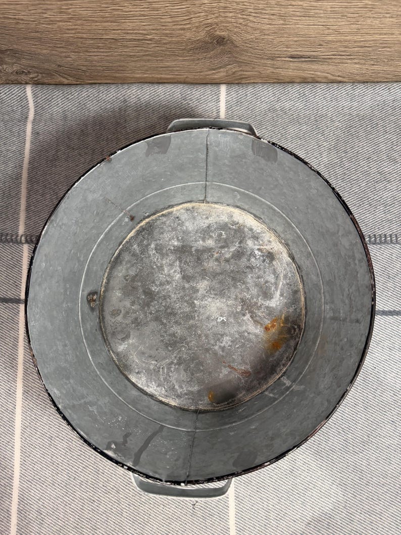 Op de afbeelding: Een ronde bak van gegalvaniseerd metaal met een verweerde, grijze afwerking en zichtbare tekenen van oxidatie. De container is voorzien van twee stevige, ge&iuml;ntegreerde metalen handgrepen aan weerszijden en een gladde, ronde bodem.