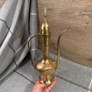 Puede incluir: Una jarra de latón vintage con un pico largo y detalles ornamentados. La jarra tiene un cuerpo bulboso y un cuello alto y esbelto con un remate decorativo. El asa se curva elegantemente de arriba a abajo. La jarra se sostiene contra una manta a cuadros gris y blanca.