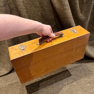 Op de afbeelding: Een rechthoekige houten kist met een bruin lederen handvat en metalen sluitingen. De kist heeft een lichtbruine afwerking met zichtbare houtnerf en enkele krassen. De handgreep is bevestigd met metalen hardware.