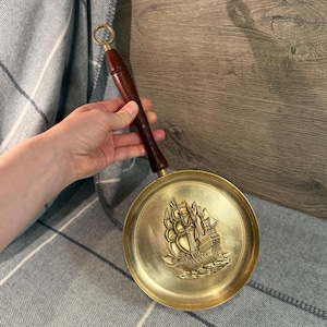 May include: A brass-colored, round pan with a dark wooden handle and a brass ring. The pan features a detailed embossed image of a sailing ship. The handle is dark brown and the pan is held by a person.